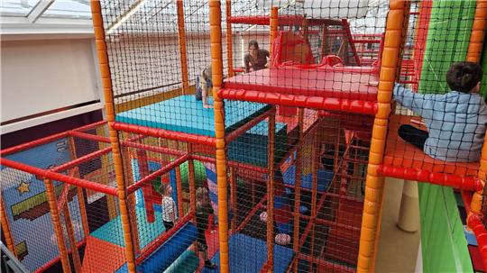 Mehrstöckige Kletteranlage mit Netzumzäunung und buntem Bodenbelag in einem Innenraum mit Kindern, die klettern und spielen.