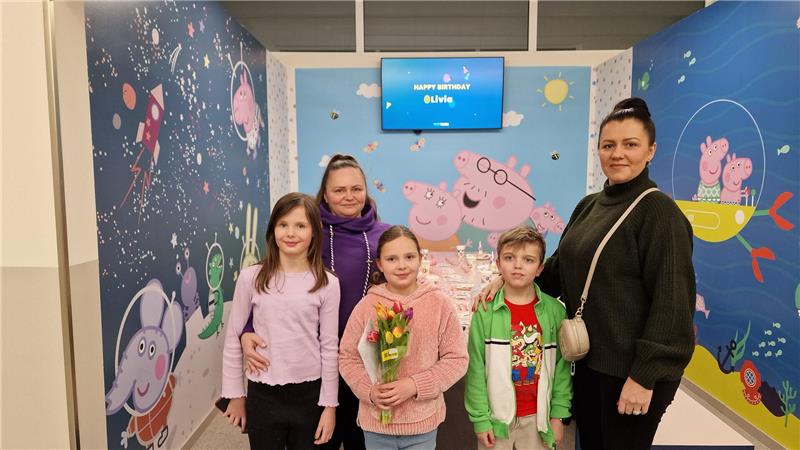 Fünf Personen stehen vor einer Wand mit Peppa-Wutz-Motiven und einem Bildschirm mit der Aufschrift „HAPPY BIRTHDAY Olivia“.