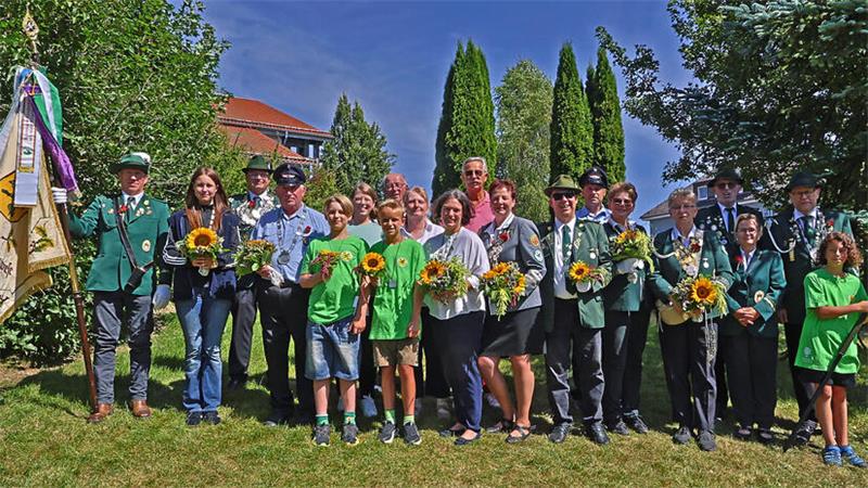 Gruppe von Menschen, darunter Kinder und Erwachsene, einige in Uniform mit Mützen, andere in grünen T-Shirts, die Sonnenblumen halten, stehen auf einer Wiese vor Büschen und Bäumen. 
