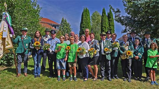 Gruppe von Menschen, darunter Kinder und Erwachsene, einige in Uniform mit Mützen, andere in grünen T-Shirts, die Sonnenblumen halten, stehen auf einer Wiese vor Büschen und Bäumen. 