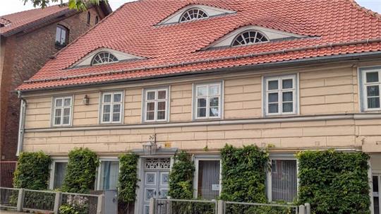 Zweistöckiges Haus mit rotem Ziegeldach, mehreren Fenstern und einer weißen Eingangstür, umgeben von grünem Bewuchs und einem niedrigen Zaun