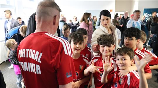 Mehrere Kinder in roten Trikots stehen in einer Gruppe mit einem Erwachsenen in rotem Shirt mit der Aufschrift 'PORTUGAL TRAINER' auf dem Rücken in einem Innenraum mit weiteren Personen im Hintergrund.