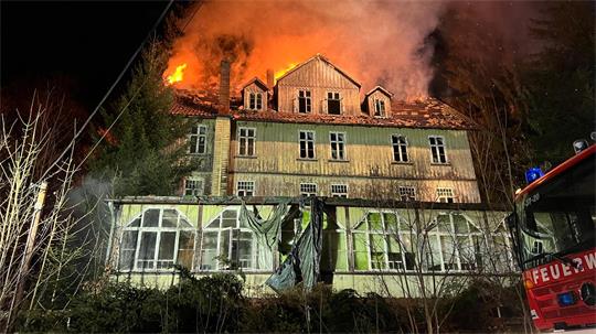 Mehrstöckiges Holzhaus bei Nacht mit brennendem Dach und Feuerwehrfahrzeug am rechten Bildrand