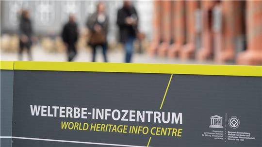 Schwarzes Schild mit gelbem Rand und weiß-gelber Aufschrift 'WELTERBE-INFOZENTRUM WORLD HERITAGE INFO CENTRE' vor unscharfem Hintergrund mit Personen und Gebäuden