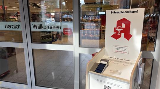 Eingangstür eines Geschäfts mit den Worten 'Herzlich Willkommen' auf den Glastüren und einem Aufsteller mit dem Text 'E-Rezepte einlösen!' und einem roten Apotheken-Logo
