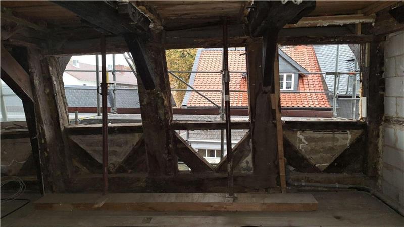 Blick durch ein altes, teilweise renovierungsbedürftiges Dachgeschoss mit sichtbaren Holzbalken und Fenstern, durch die Häuserdächer zu sehen sind.