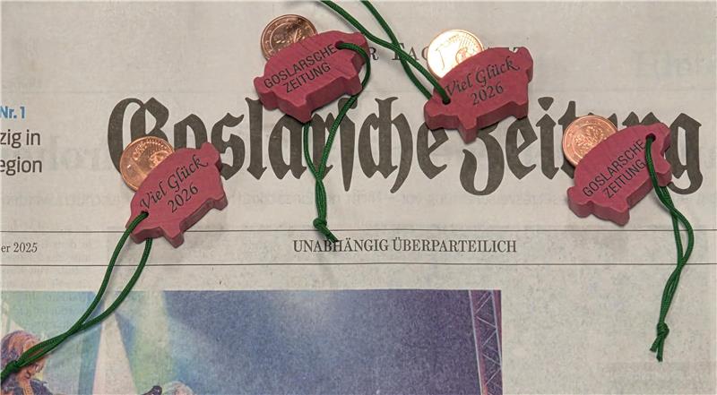 Zeitung mit der Überschrift 'Goslarsche Zeitung', darauf liegen vier kleine rosafarbene Schweinchen-Anhänger mit der Aufschrift 'Goslarsche Zeitung' und 'Viel Glück 2026'