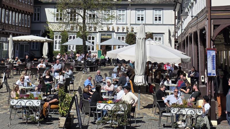 Belebter Außenbereich eines Cafés mit mehreren Tischen, Sonnenschirmen und Fachwerkhäusern im Hintergrund.