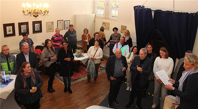Gruppe von Menschen in einem Raum mit Holzboden und Kronleuchter, einige halten Papiere oder Getränke, an den Wänden hängen gerahmte Bilder