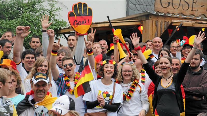 Menschenmenge mit erhobenen Armen, einige tragen schwarz-rot-goldene Accessoires und halten Deutschlandfahnen, eine Person hält ein rotes Handschild mit der Aufschrift 'Goal!'