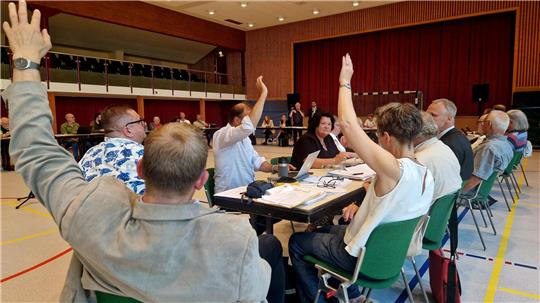 Ratsleute sitzen in der Stadthalle und manche von ihnen heben die Hand.