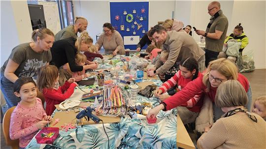 Kinder basteln an einem bunten Tisch, Erwachsene beugen sich zu ihnen herunter und helfen ihnen.