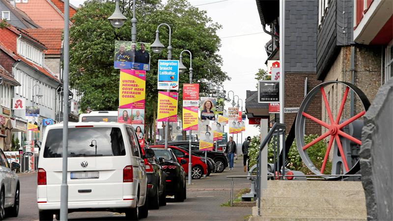 Straßenszene mit parkenden Autos, mehreren Werbeplakaten an Laternen und einem großen Rad auf einem Sockel am rechten Bildrand
