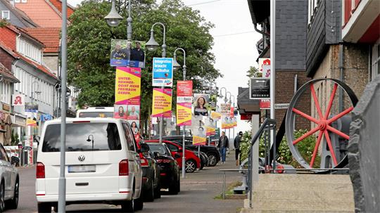 Straßenszene mit parkenden Autos, mehreren Werbeplakaten an Laternen und einem großen Rad auf einem Sockel am rechten Bildrand