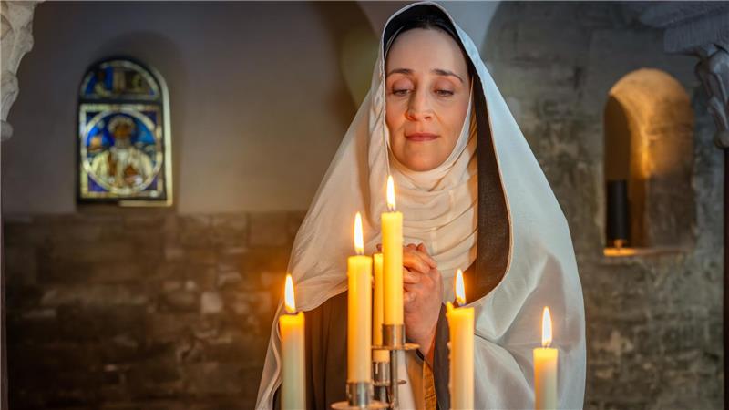 Person in weißem Gewand mit Kapuze steht hinter einem Kerzenleuchter mit brennenden Kerzen in einem Raum mit Steinwänden und einem bunten Kirchenfenster