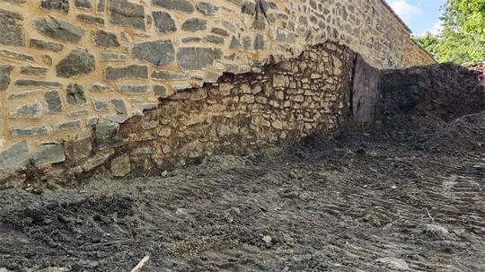 Mauer mit teilweise freigelegtem, älterem Mauerwerk und davor unebener, erdiger Boden