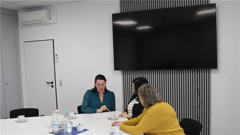 Drei Personen sitzen an einem weißen Konferenztisch in einem modernen Besprechungsraum mit großem Flachbildschirm an der Wand.