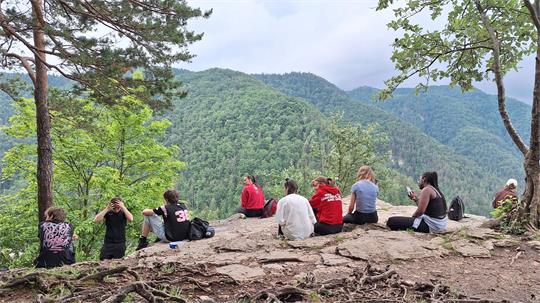 Gruppe von Menschen sitzt auf einem Felsen und blickt auf bewaldete Berge.