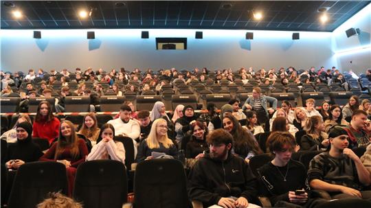 Große Gruppe von Menschen sitzt in einem Kinosaal mit mehreren Sitzreihen und dunklen Polstersesseln.