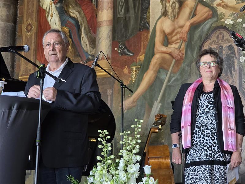 Zwei Personen stehen vor einer Wand mit einem großen Wandgemälde, ein Mann spricht an einem Mikrofon, eine Frau trägt ein gemustertes Kleid und einen rosa Schal