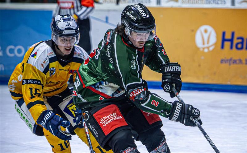 Zwei Eishockeyspieler in Aktion, einer in grün-schwarzer Ausrüstung mit Puck, der andere in gelb-schwarzer Ausrüstung verfolgt ihn auf dem Eis.