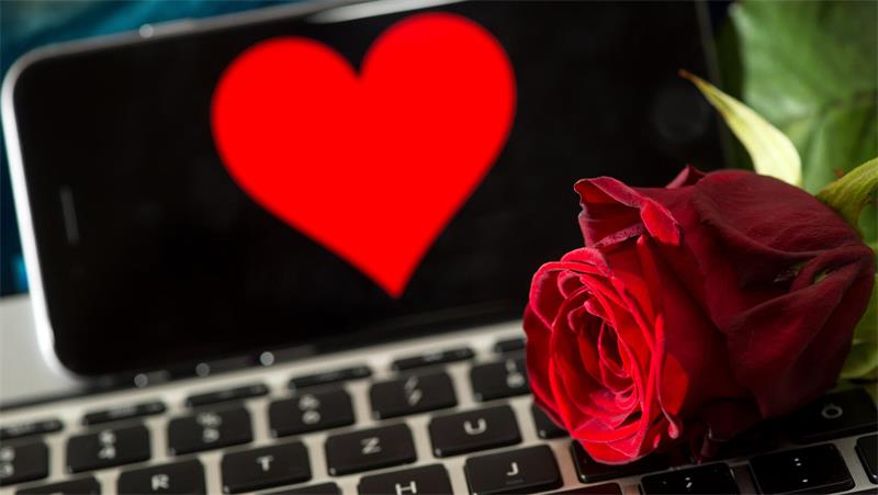 Auf einer Computertastatur liegt eine rote Rose, dahinter ist ein Smartphone zu sehen, auf dessen Bildschirm ein rotes Herz erscheint.