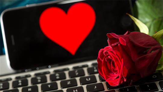 Auf einer Computertastatur liegt eine rote Rose, dahinter ist ein Smartphone zu sehen, auf dessen Bildschirm ein rotes Herz erscheint.