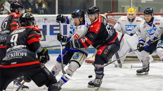 Eishockeyspieler in Aktion vor dem Tor, zwei Spieler kämpfen um den Puck, Torwart und weitere Spieler im Hintergrund