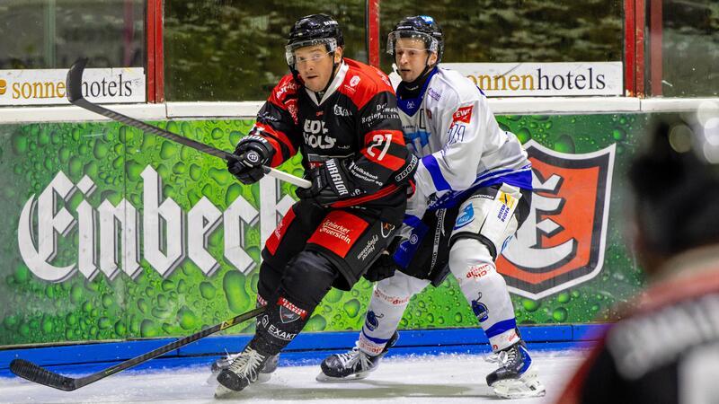 Zwei Eishockeyspieler in Aktion auf dem Eis, einer in schwarzer und roter, der andere in weißer Ausrüstung, im Hintergrund Werbetafeln