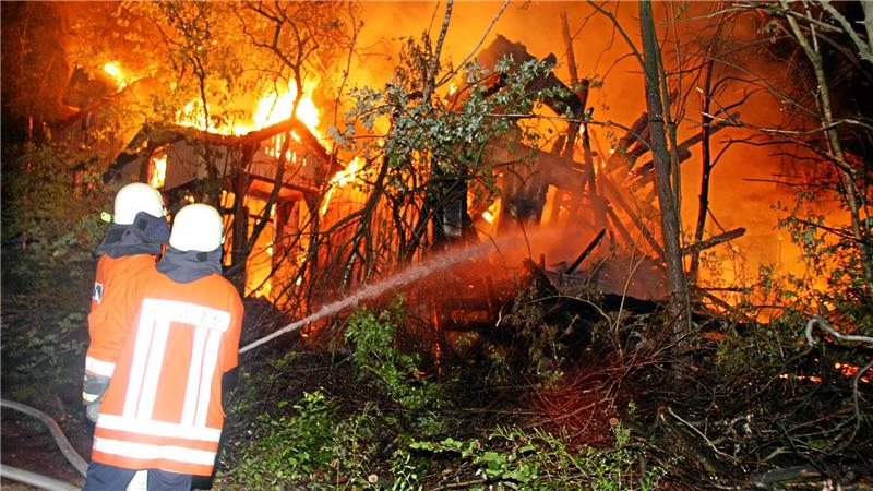 Zwei Feuerwehrleute in Schutzkleidung löschen mit einem Schlauch ein Feuer in einem Waldgebiet mit brennenden Bäumen und Gebäuderesten