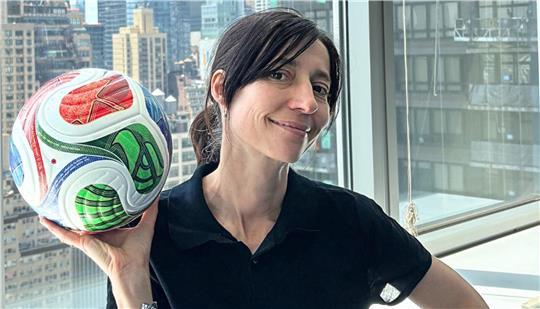 Das Foto zeigt Lisa Hollenbach vom Generalkonsulat der Bundesrepublik Deutschland in Manhattan mit einem Fußball in der rechten Hand. Aus dem Fenster hinter ihr ist die Skyline von Manhattan zu sehen.