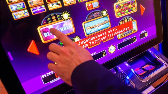 Hand zeigt auf einen Touchscreen eines Spielautomaten mit mehreren Spieloptionen und dem Hinweis 'Jugendschutz aktiv am Terminal freischalten'