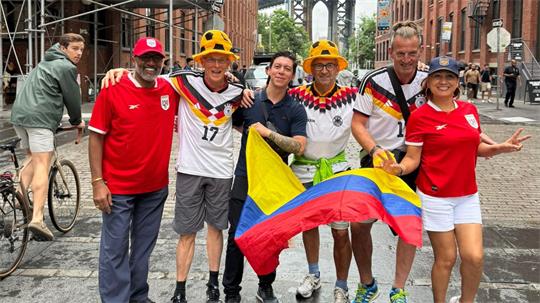 Gruppe von sechs Personen in Fußballtrikots verschiedener Länder posiert auf gepflasterter Straße, eine Person hält eine kolumbianische Flagge