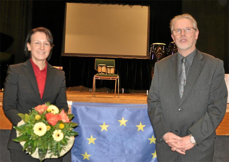 Zwei Personen in formeller Kleidung stehen vor einer Bühne mit einem Tisch, der mit einer Europaflagge bedeckt ist, eine Person hält einen Blumenstrauß