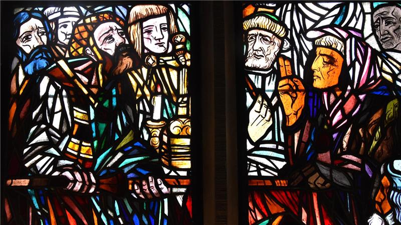 Buntes Kirchenfenster mit mehreren sitzenden Figuren in mittelalterlicher Kleidung und Kerzen auf einem Tisch