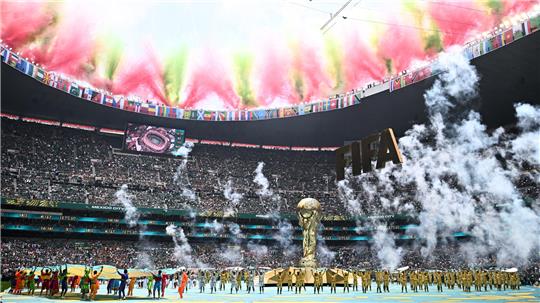 Blick in ein vollbesetztes Stadion mit einer großen goldenen FIFA-Weltmeisterschaftstrophäe in der Mitte und buntem Rauch über den Zuschauerrängen.