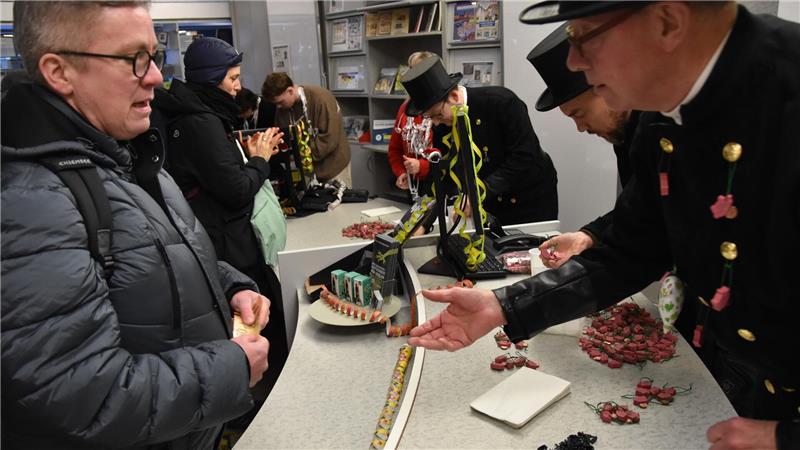 Mehrere Personen stehen an einem Tisch mit verschiedenen handwerklichen oder künstlerischen Objekten, darunter eine lange, bunte Kette und kleine rote Figuren