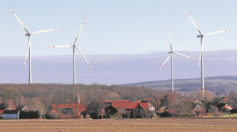 Hinter einem Dorf erheben sich vier Windenergieanlagen.