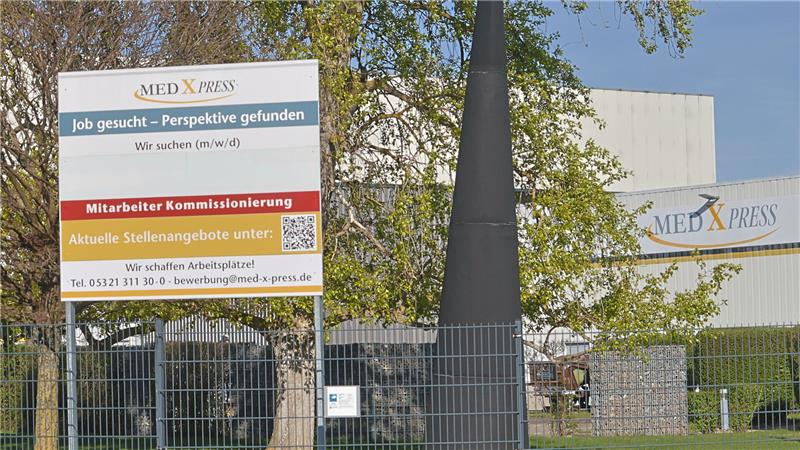 Firmengelände mit einem großen Werbeschild für Stellenangebote der Firma MED X PRESS vor einem Zaun und Bäumen