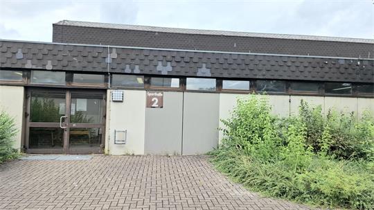 Eingang eines Gebäudes mit zwei großen grauen Türen, links davon ein Glaseingang mit braunem Rahmen und davor gepflasterter Boden sowie rechts und links Sträucher