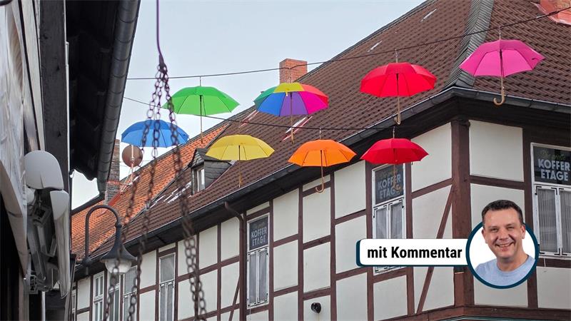 Bunte Regenschirme hängen über einer Fachwerkhausfassade mit einem Geschäft im Erdgeschoss und Blumen im Vordergrund