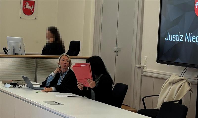 Zwei Personen sitzen an einem langen weißen Tisch in einem hellen Raum mit großen Fenstern und einem Bildschirm mit dem Schriftzug 'Justiz Niedersachsen'