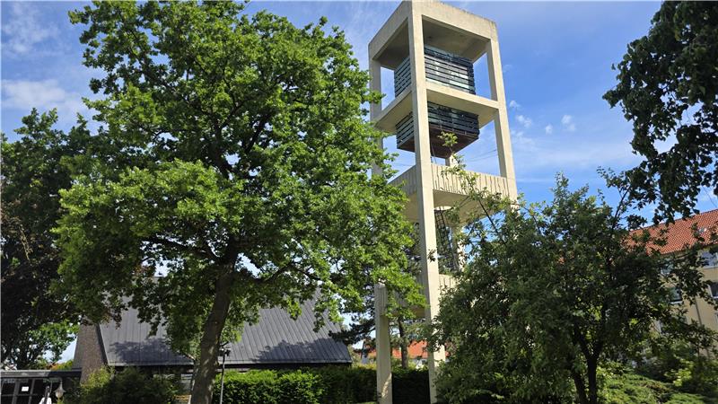 Moderner Kirchturm mit offenem Glockenstuhl umgeben von Bäumen und Gebäuden unter blauem Himmel.