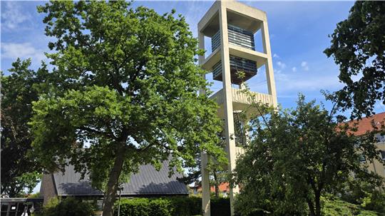 Moderner Kirchturm mit offenem Glockenstuhl umgeben von Bäumen und Gebäuden unter blauem Himmel.