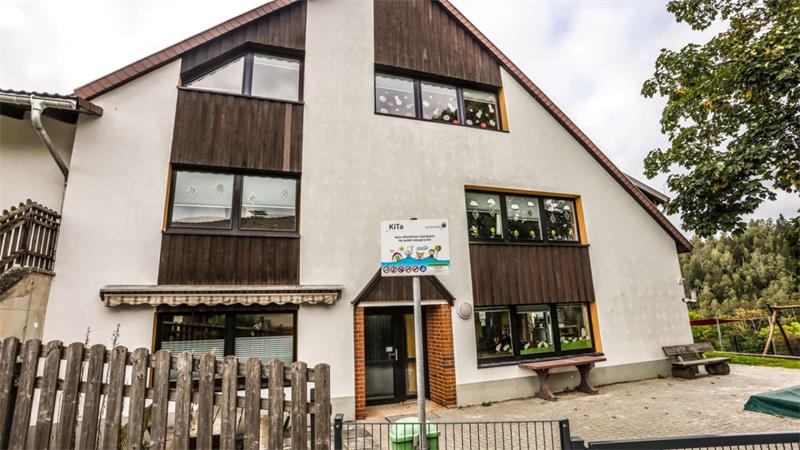 Mehrstöckiges Wohnhaus mit weißer Fassade und braunen Holzverkleidungen, davor ein Zaun und ein Schild vor dem Eingang