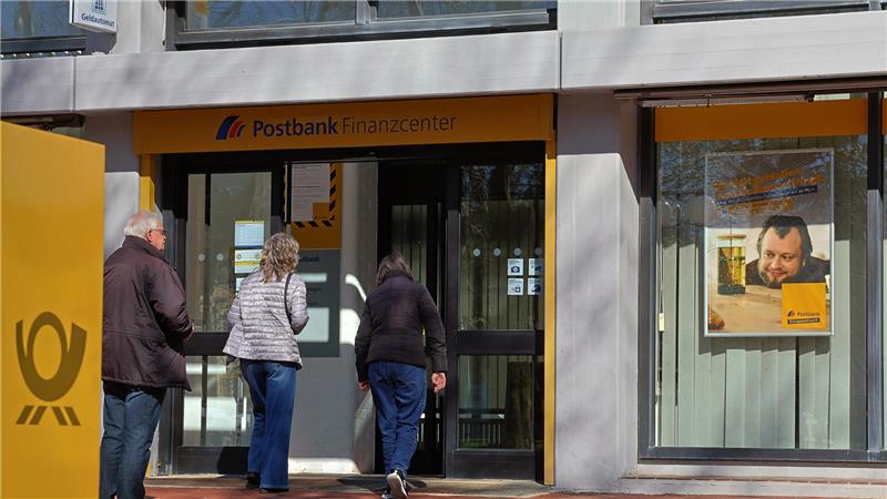 Drei Personen stehen vor dem Eingang eines Postbank-Finanzcenters mit gelber Fassade und Glasfront.