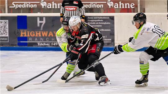 Drei Eishockeyspieler kämpfen um den Puck auf dem Eis, im Hintergrund steht ein Schiedsrichter vor einer Werbewand