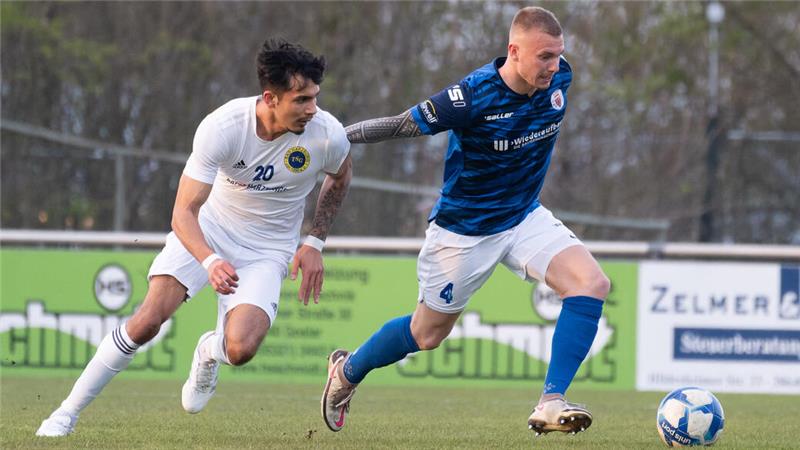 Zwei Fußballspieler in Aktion, einer in weißem Trikot mit der Nummer 20, der andere in blauem Trikot mit der Nummer 4, beide laufen auf einen Fußball zu.
