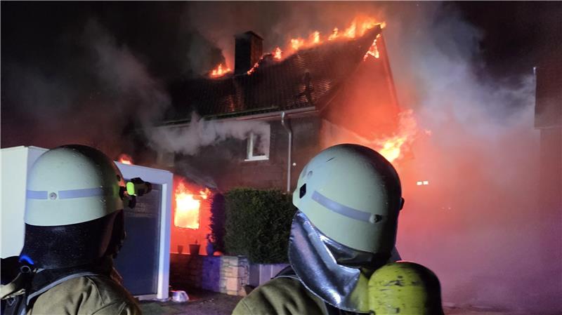 Zwei Feuerwehrleute stehen mit Sauerstoffflaschen und Ausrüstung vor einem brennenden Haus.