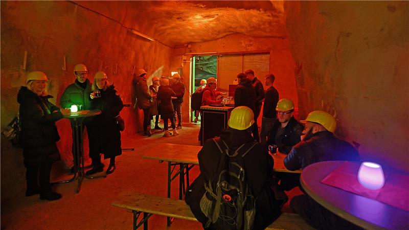 Großer Besucheransturm auf Goslars Weihnachtsmarkt unter Tage Ein mit orangefarbenem Licht ausgestrahlter Raum unter Tage ist mit Stehtischen und Bierzeltgarnitur möbliert. Menschen mit gelben Helmen stehen und sitzen bei einem Getränk zusammen.